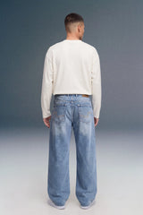 A man viewed from behind wearing a cream long-sleeve shirt and light-wash blue wide-leg jeans against a dark gray studio background.