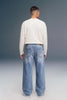 A man viewed from behind wearing a cream long-sleeve shirt and light-wash blue wide-leg jeans against a dark gray studio background.