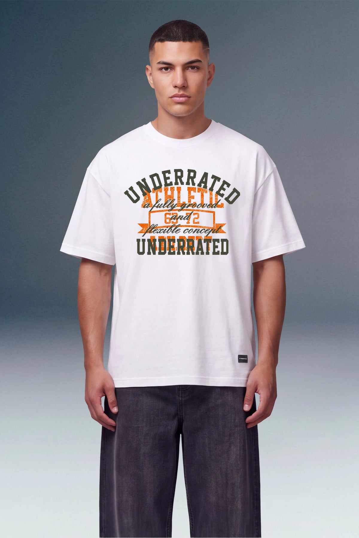 A male model wearing a white graphic t-shirt with 'UNDERRATED ACHIEVED' printed in dark green and orange, paired with dark gray pants, against a studio background.