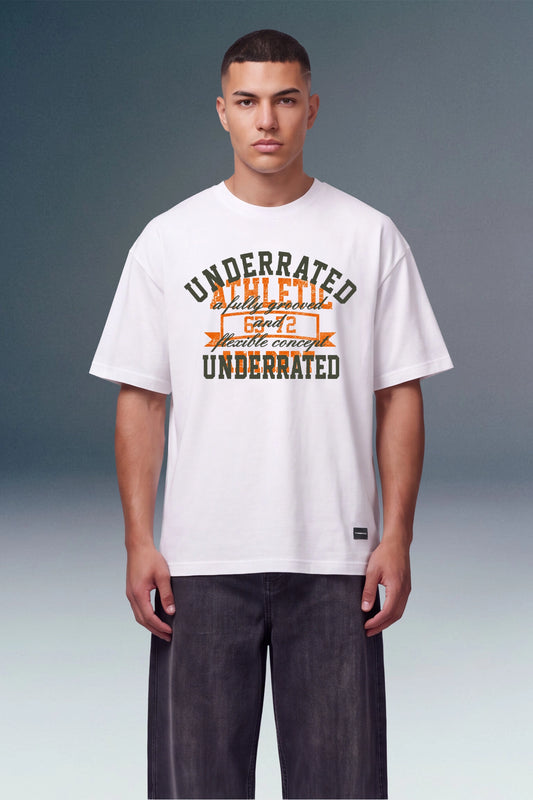 A male model wearing a white graphic t-shirt with 'UNDERRATED ACHIEVED' printed in dark green and orange, paired with dark gray pants, against a studio background.