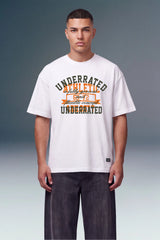 A male model wearing a white graphic t-shirt with 'UNDERRATED ACHIEVED' printed in dark green and orange, paired with dark gray pants, against a studio background.