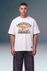 A male model wearing a white graphic t-shirt with 'UNDERRATED ACHIEVED' printed in dark green and orange, paired with dark gray pants, against a studio background.