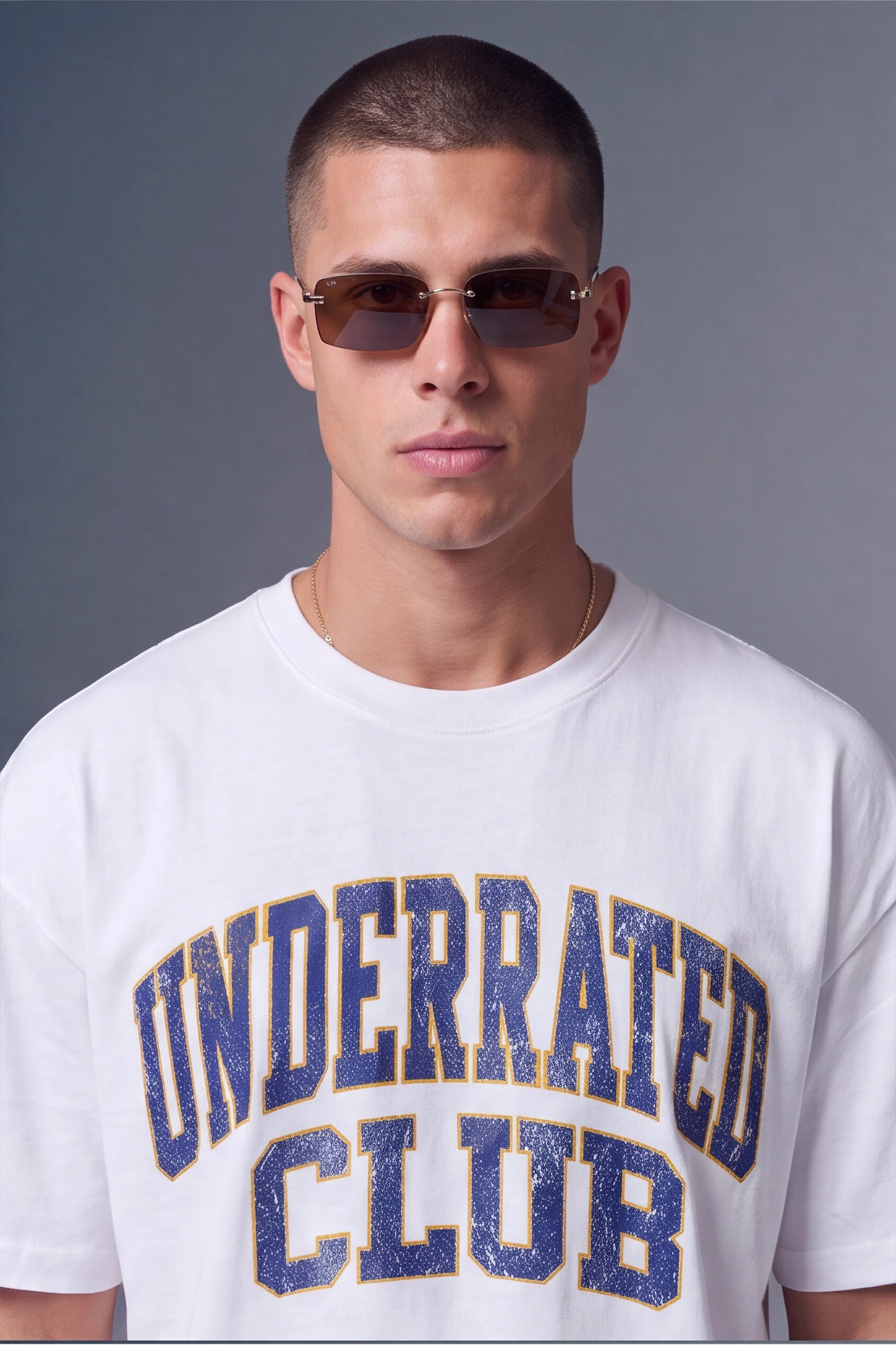 A man wearing a white crewneck t-shirt with 'UNDERRATED CLUB' printed in distressed blue varsity lettering with gold outlines, paired with dark sunglasses, against a solid gray studio background.