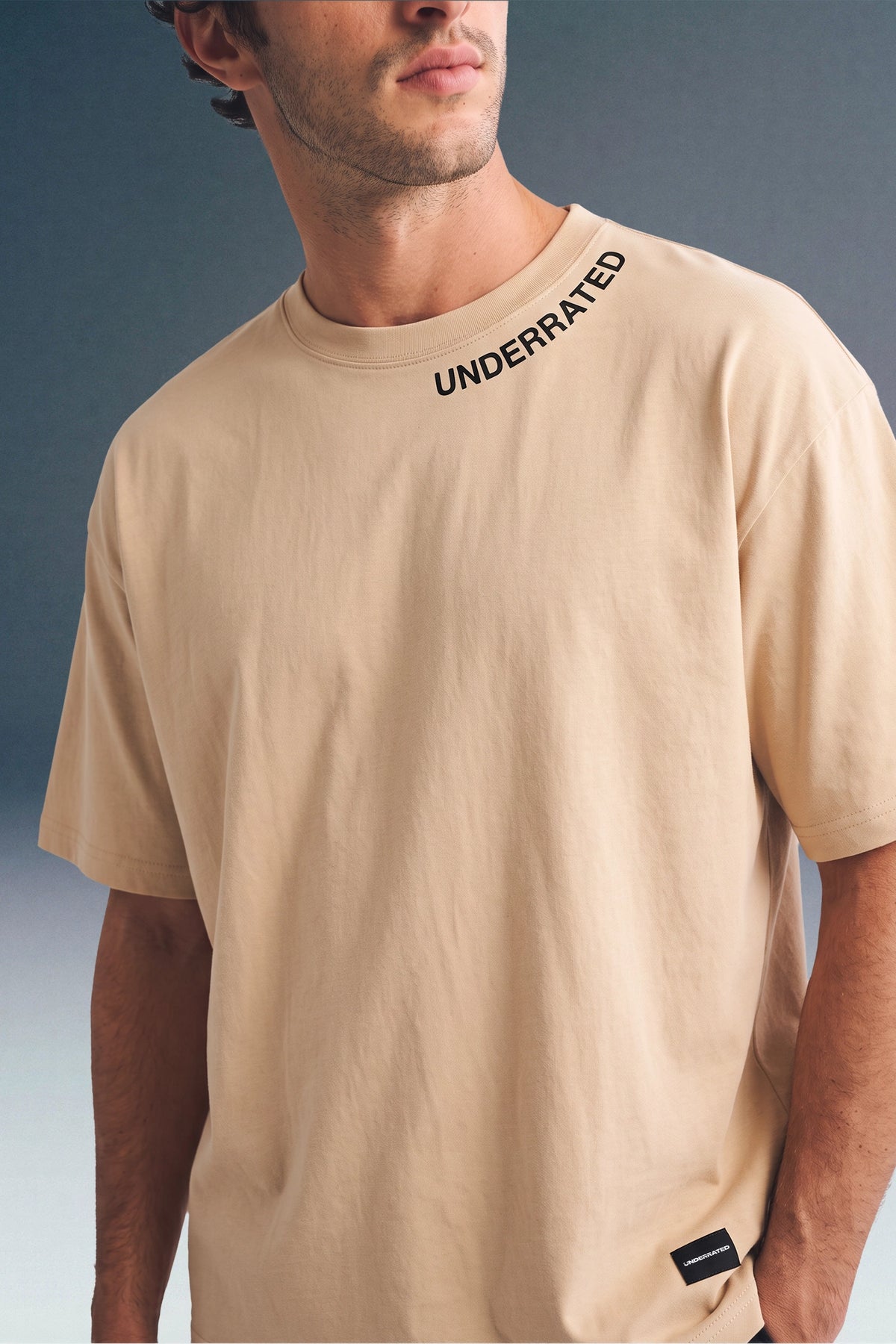 Medium close-up shot from the chest up of a man wearing a beige T-shirt with black text along the collar seam and a horizontal seam across the chest.