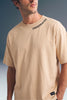 Medium close-up shot from the chest up of a man wearing a beige T-shirt with black text along the collar seam and a horizontal seam across the chest.