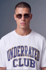 A man wearing a white crewneck t-shirt with 'UNDERRATED CLUB' printed in distressed blue varsity lettering with gold outlines, paired with dark sunglasses, against a solid gray studio background.