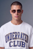A man wearing a white crewneck t-shirt with 'UNDERRATED CLUB' printed in distressed blue varsity lettering with gold outlines, paired with dark sunglasses, against a solid gray studio background.