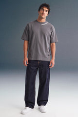 Full-length front view of a man standing straight against a grey studio backdrop, wearing a dark grey T-shirt, loose dark navy trousers, and white sneakers.
