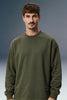 A man wearing an olive green crewneck sweatshirt, photographed against a dark blue-gray studio background.