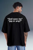 A man viewed from the back wearing a black oversized t-shirt with white text that reads “God save me” “jpg or png?” and blue jeans, set against a gray studio background.