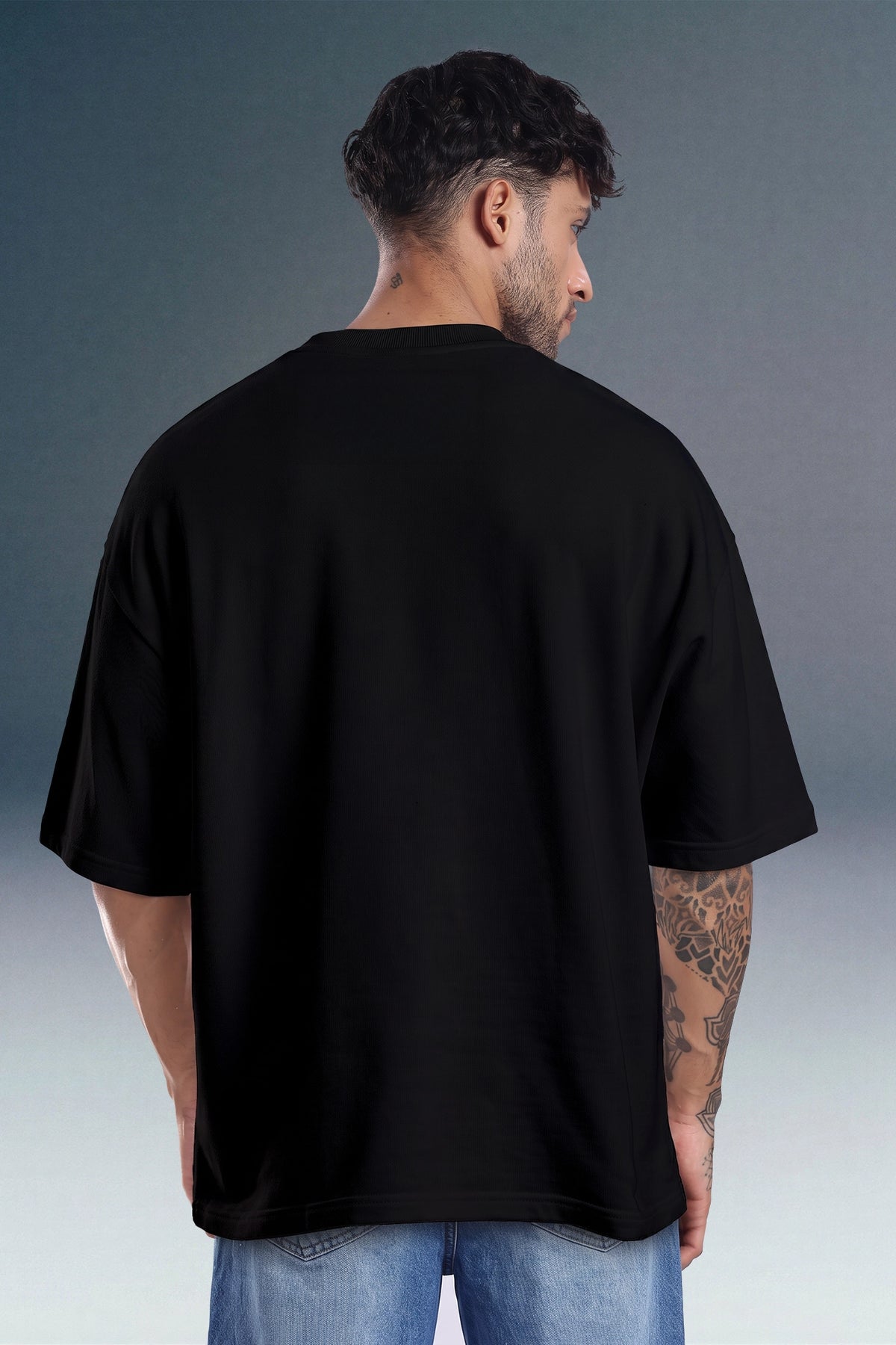 A man viewed from behind wearing an oversized black short-sleeve t-shirt and blue jeans, standing against a dark gray studio background.