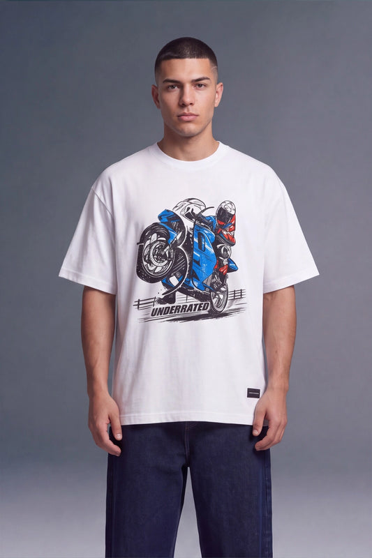 A man wearing a white graphic t-shirt with a blue and black motorcycle print and the text 'UNDERRATED' on the front, paired with dark blue jeans, against a gray studio background.