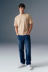 Man wearing a beige t-shirt, blue jeans, and white sneakers against a gray background