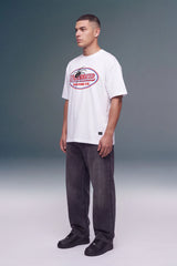 A man wearing a white t-shirt with a red and black 'Unleashed Racing Co' graphic, paired with dark gray jeans and black sneakers, standing against a gray studio background.