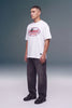 A man wearing a white t-shirt with a red and black 'Unleashed Racing Co' graphic, paired with dark gray jeans and black sneakers, standing against a gray studio background.