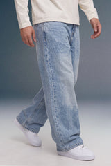 A person wearing light-wash, wide-leg denim jeans with a faded wash, styled with a cream long-sleeve shirt and white sneakers against a studio backdrop.