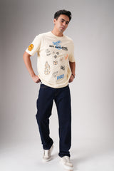 Man posing casually in beige graphic t‑shirt and dark jeans with hands in pockets against grey studio background.
