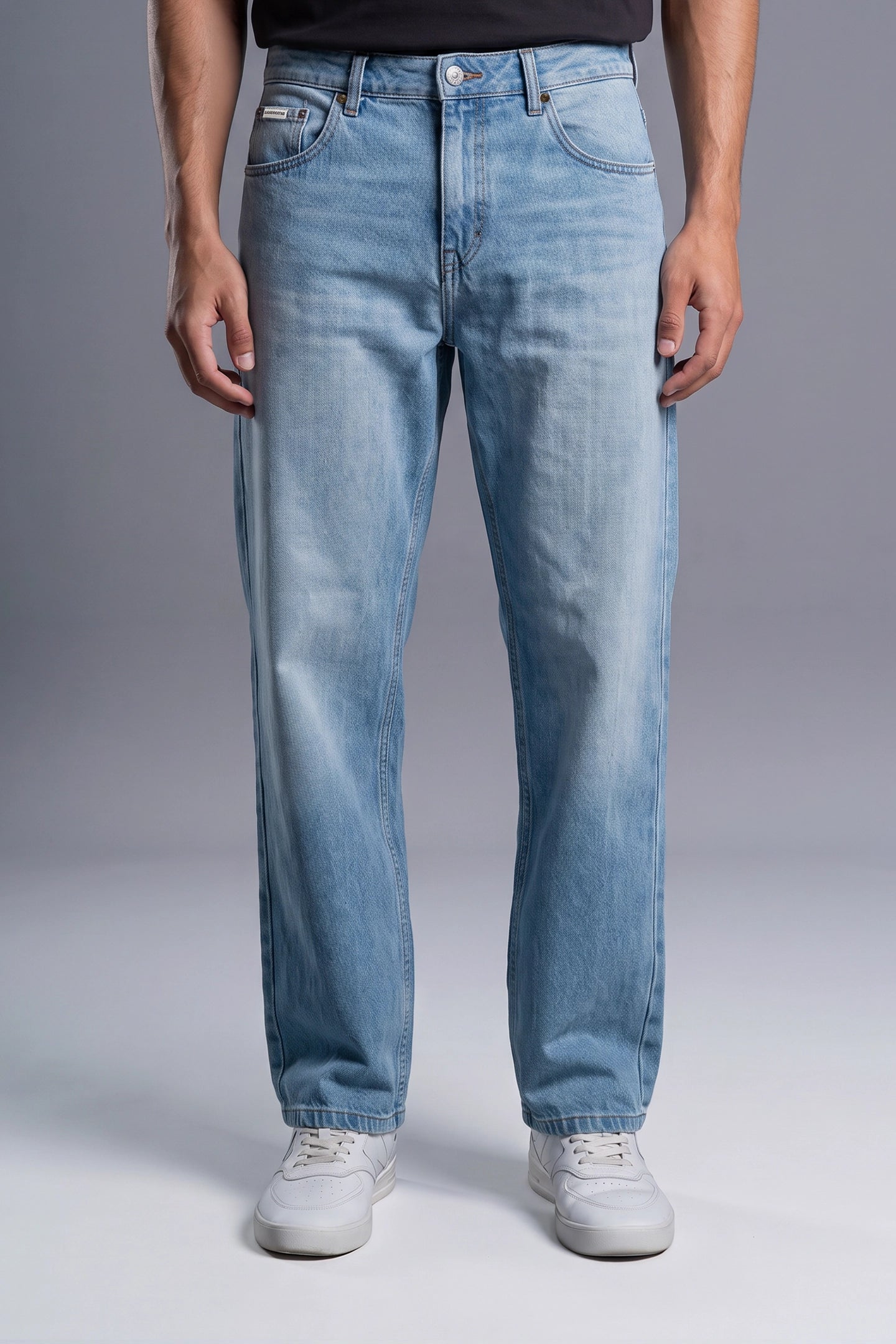 A person wearing light blue straight-leg denim jeans with a faded wash, paired with a black top and white sneakers, shown from the waist down against a gray studio background.