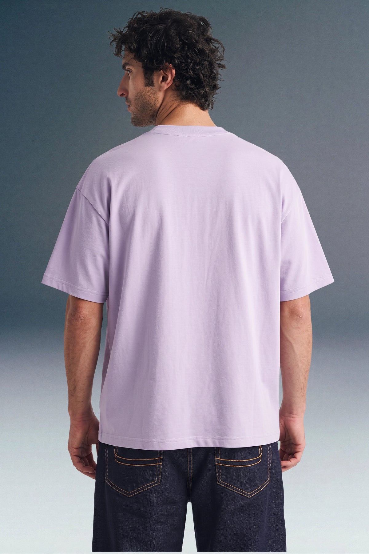 Rear view from the waist up of a man looking away, showing the back profile and relaxed sleeve length of a light purple T-shirt paired with dark jeans.