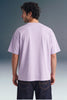 Rear view from the waist up of a man looking away, showing the back profile and relaxed sleeve length of a light purple T-shirt paired with dark jeans.