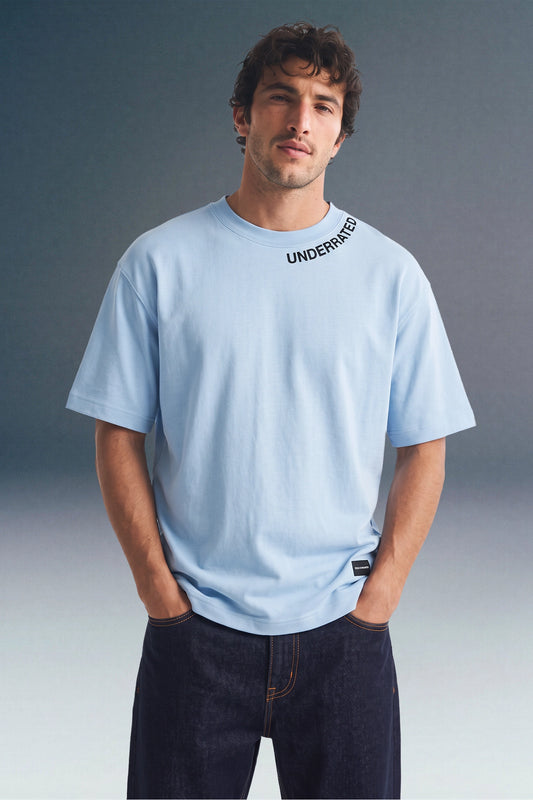 Here is the image alt text for each of the 6 vertical panels in image_06d338.png, listed from top to bottom:   First image: Front view from the waist up of a man with curly hair standing with his hands in his pockets, wearing a light blue crewneck T-shirt featuring subtle black collar text along the neckline.