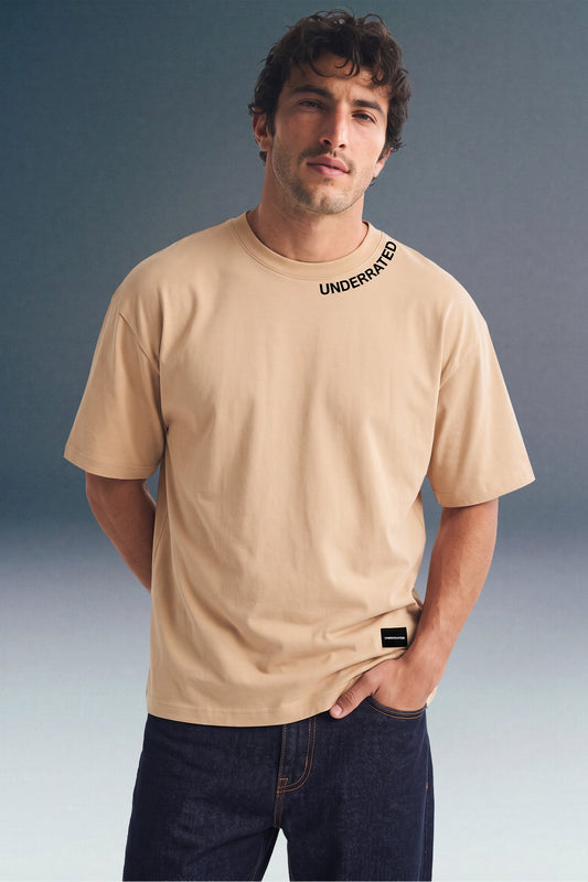 Front view from the waist up of a man looking forward with one hand in his pocket, wearing a beige crewneck T-shirt with black print along the left side of the collar.