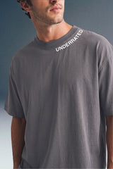 Medium close-up shot from the chest up of a man looking to the side, wearing a dark grey crewneck T-shirt with small white text printed along the left side of the collar.