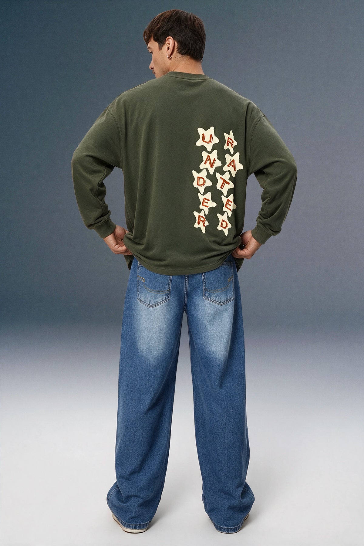 A man viewed from behind wearing an olive green long-sleeve sweatshirt with a vertical graphic of cream and red stars on the back, paired with loose blue jeans, against a studio backdrop.