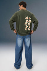 A man viewed from behind wearing an olive green long-sleeve sweatshirt with a vertical graphic of cream and red stars on the back, paired with loose blue jeans, against a studio backdrop.
