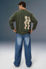 A man viewed from behind wearing an olive green long-sleeve sweatshirt with a vertical graphic of cream and red stars on the back, paired with loose blue jeans, against a studio backdrop.