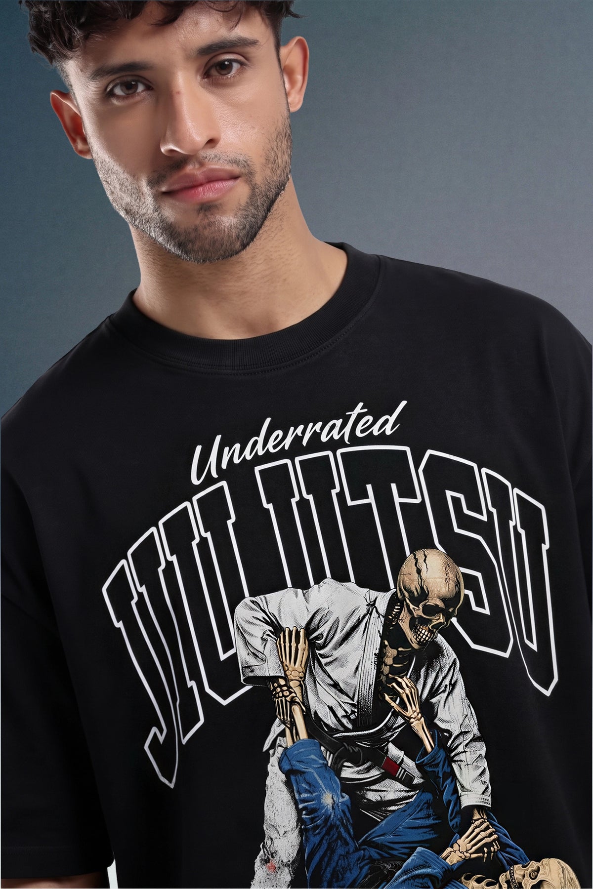 A man wearing a black graphic t-shirt with 'Underrated Jiu Jitsu' and a skeleton fighting illustration on the front, set against a dark gray studio background.