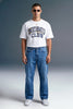 A man wearing a white graphic t-shirt with 'UNDERRATED CLUB' printed in navy blue, paired with blue jeans and white sneakers, standing against a gray studio background.