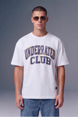 A man wearing a white crewneck t-shirt with 'UNDERRATED CLUB' printed in navy blue distressed lettering across the chest, paired with blue jeans, set against a dark gray studio background.