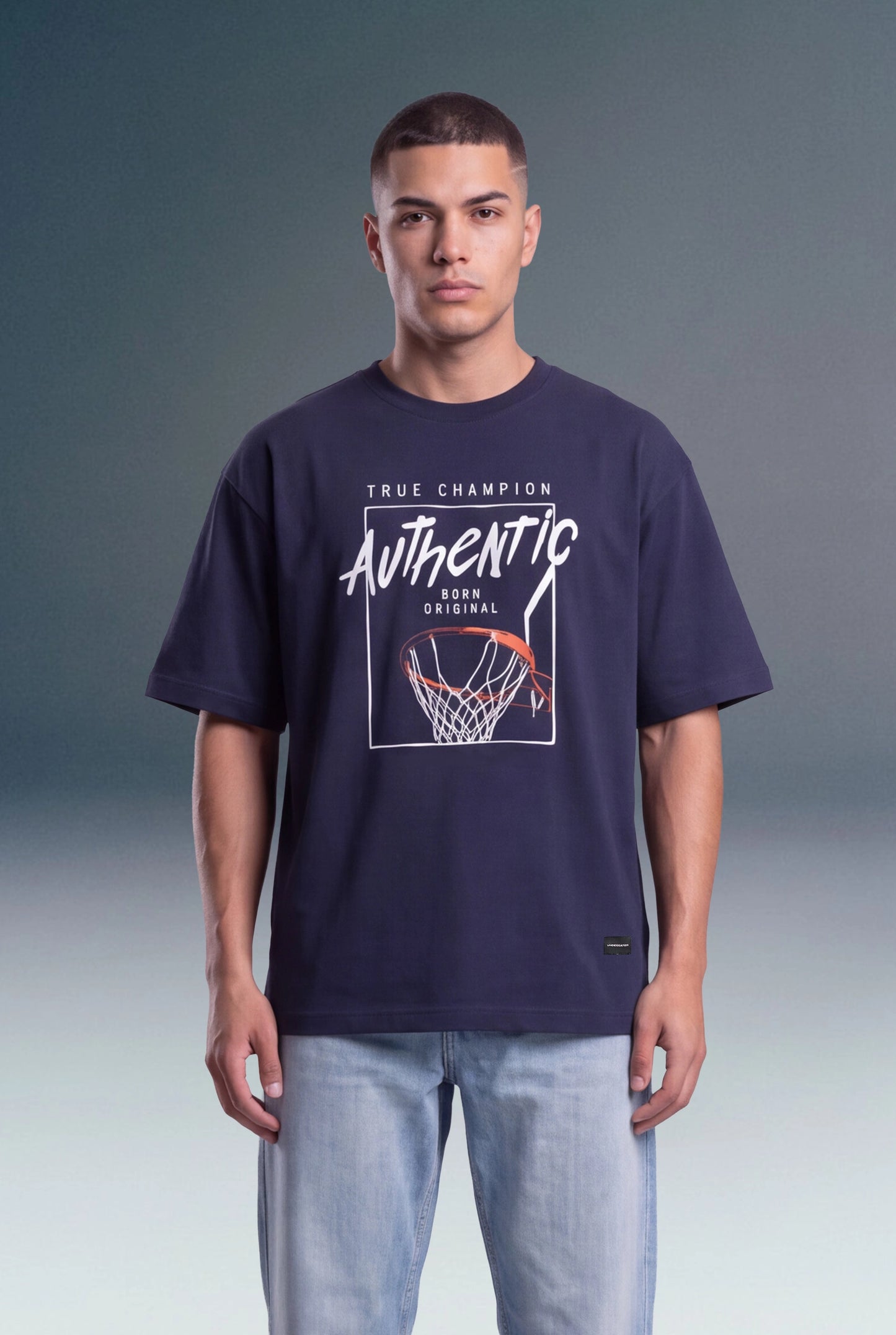A man wearing a navy blue graphic t-shirt with a basketball hoop design and the words 'True Champion Authentic' on the front, paired with light blue jeans, standing against a gray studio background.