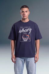 A man wearing a navy blue graphic t-shirt with a basketball hoop design and the words 'True Champion Authentic' on the front, paired with light blue jeans, standing against a gray studio background.