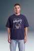 A man wearing a navy blue graphic t-shirt with a basketball hoop design and the words 'True Champion Authentic' on the front, paired with light blue jeans, standing against a gray studio background.