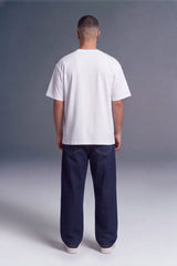 A man viewed from behind wearing a plain white short-sleeve t-shirt and dark navy blue jeans, standing against a gray studio background.