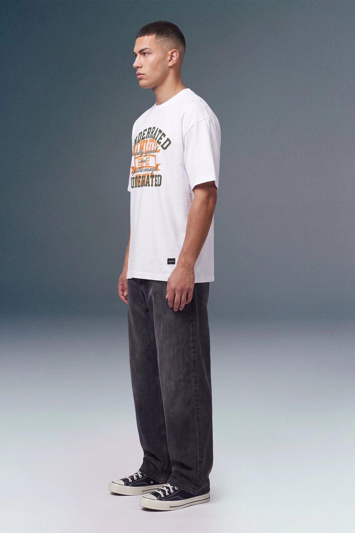 A man stands in profile wearing a white short-sleeve graphic T-shirt with orange and dark lettering, dark gray jeans, and black low-top sneakers against a smooth gray studio background.