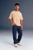 A man modeling a light beige crew-neck t-shirt and dark blue jeans, standing against a dark gray studio background.