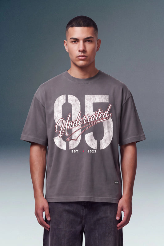 A man wearing a dark gray graphic t-shirt with a distressed white '95' print and 'Underrated' script, paired with dark gray jeans, set against a studio gradient background.