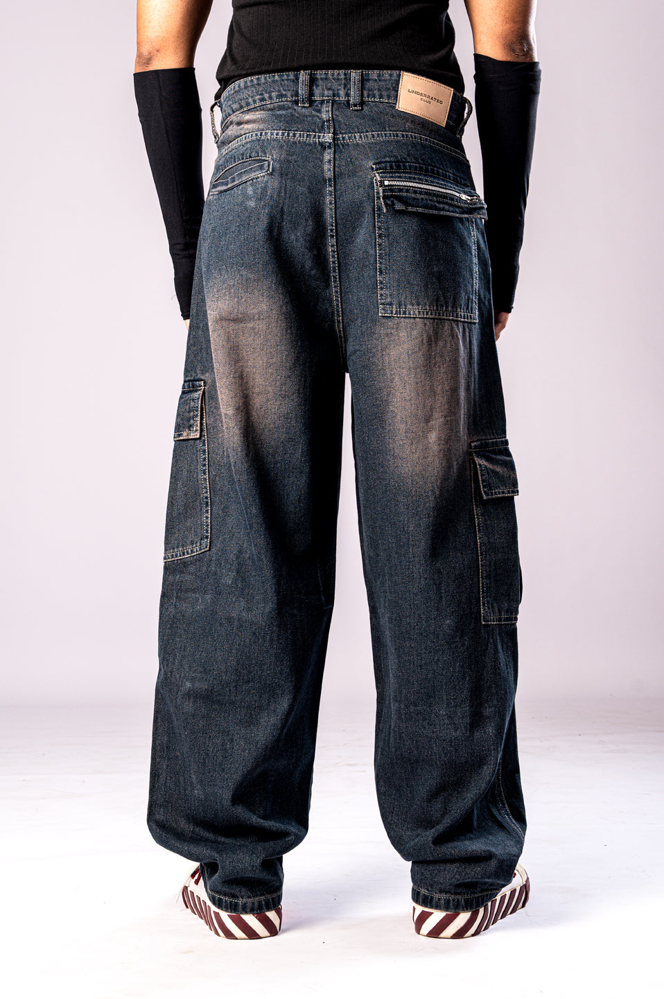 Full-body back view of a person wearing dark, rusty-blue baggy cargo jeans, a black top, and black arm sleeves, showing the loose fit and large side cargo pockets.