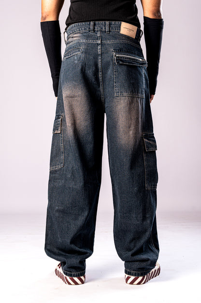 Full-body back view of a person wearing dark, rusty-blue baggy cargo jeans, a black top, and black arm sleeves, showing the loose fit and large side cargo pockets.