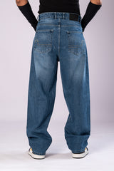 Full-body front view of a person wearing light-wash blue baggy jeans, a black top, and black arm sleeves, with their hands in their pockets.