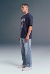 A man wearing a dark navy graphic t-shirt, light blue straight-leg jeans, and white sneakers, standing in profile against a gray studio background.