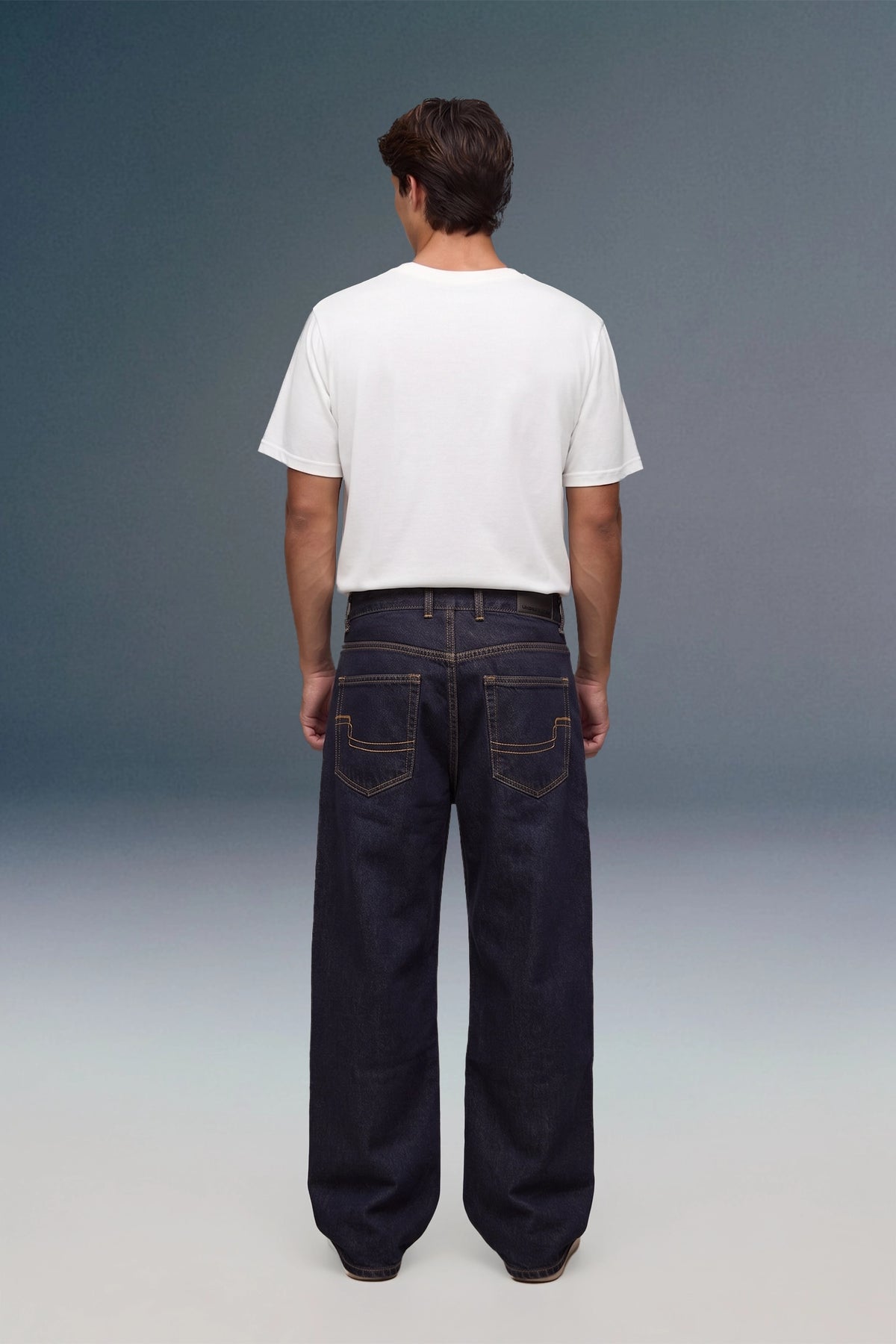 A man viewed from behind wearing a plain white short-sleeve t-shirt and dark wide-leg denim jeans, standing against a studio background with a blue-to-gray gradient.