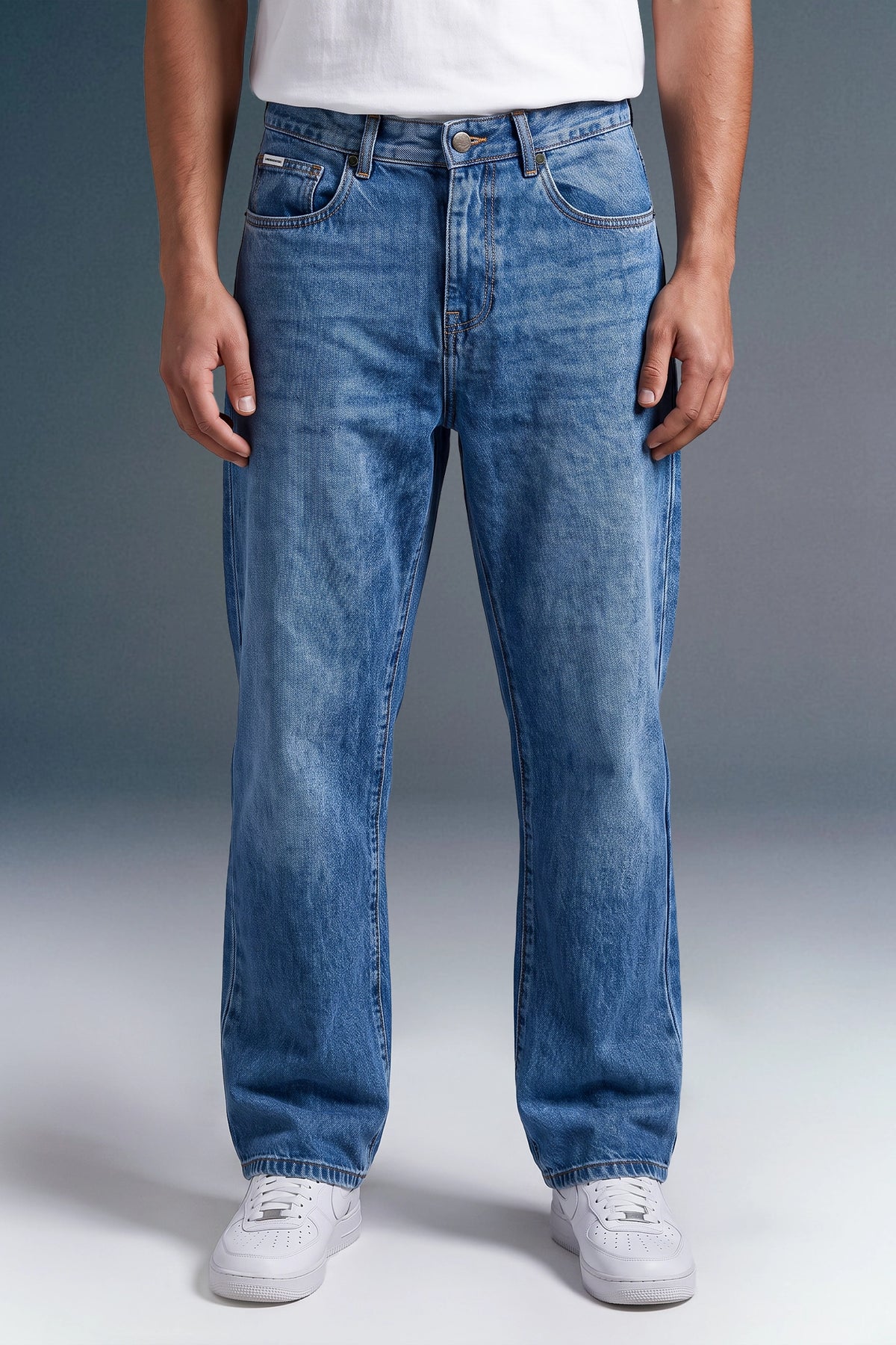 A person wearing medium-wash blue denim jeans with a white t-shirt and white sneakers, shown from the waist down against a gray studio background.