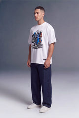 A man wearing a white graphic t-shirt with a motorcycle print, dark blue jeans, and white sneakers, standing against a gray studio background.