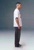 A man in profile wearing a white oversized t-shirt with a colorful graphic print on the chest, dark gray jeans, and black canvas sneakers, standing against a gray studio background.