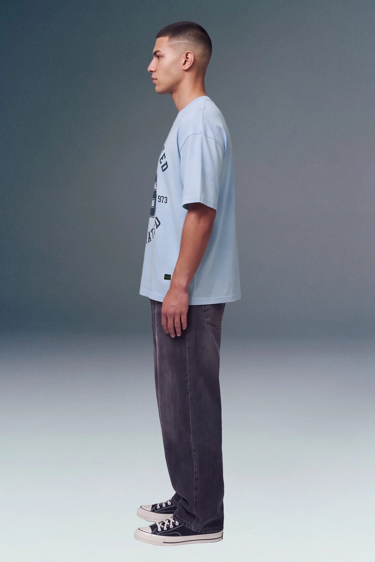 A man in profile wearing a light blue graphic t-shirt, dark gray washed jeans, and black canvas sneakers, standing against a gray studio background.
