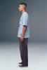 A man in profile wearing a light blue graphic t-shirt, dark gray washed jeans, and black canvas sneakers, standing against a gray studio background.
